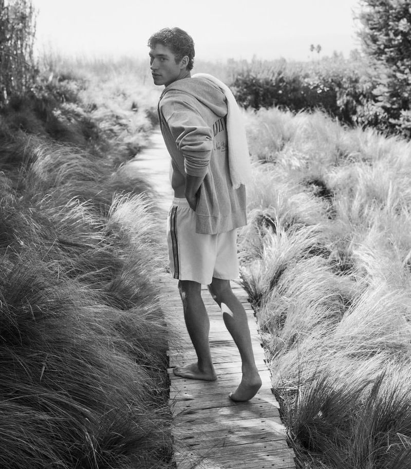 Black and white portrait of a young adult male on a wooden path in tall grass wearing shorts and a hoodie, looking over his shoulder.