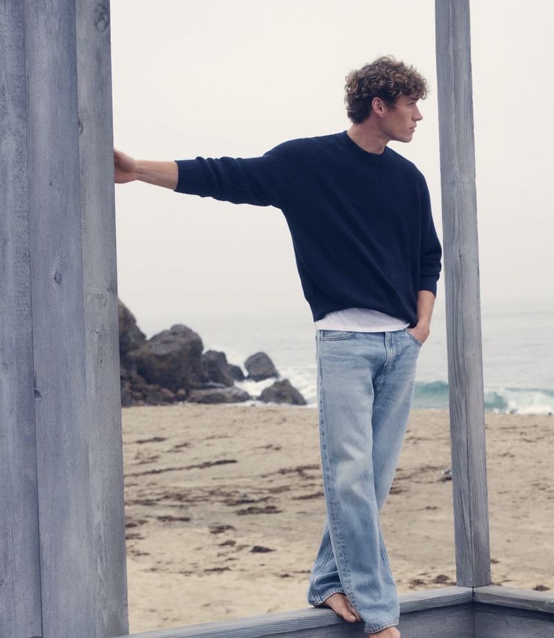 Young man leaning against a weathered wooden post on a sandy beach wearing a dark blue sweater and light blue jeans