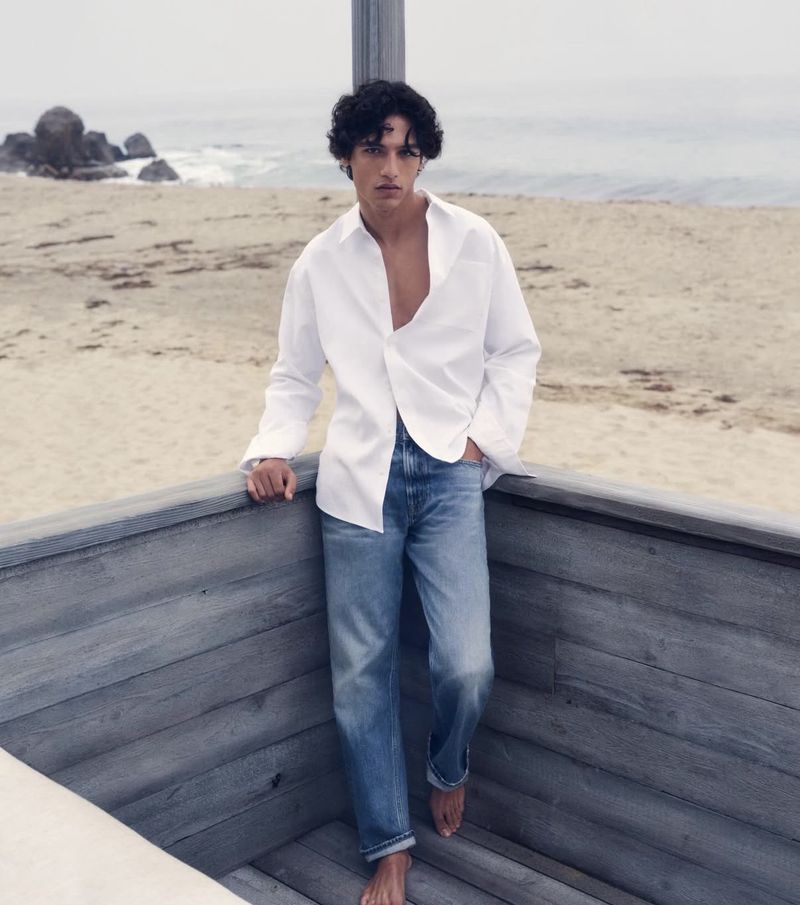 A young adult man with dark curly hair walks on a beach with a white shirt and jeans, posing on wooden steps leading to the sandy shore.
