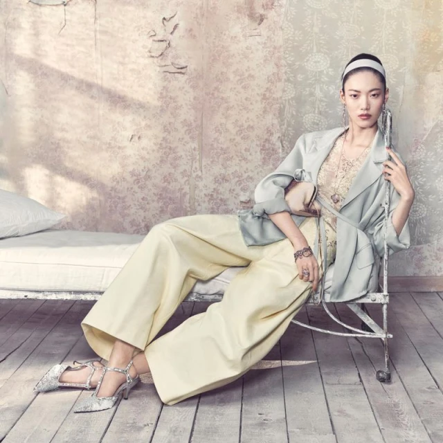 Asian woman in wide-leg pants and a blazer poses on a daybed in a vintage-style room with a patterned wall and silver high heels.