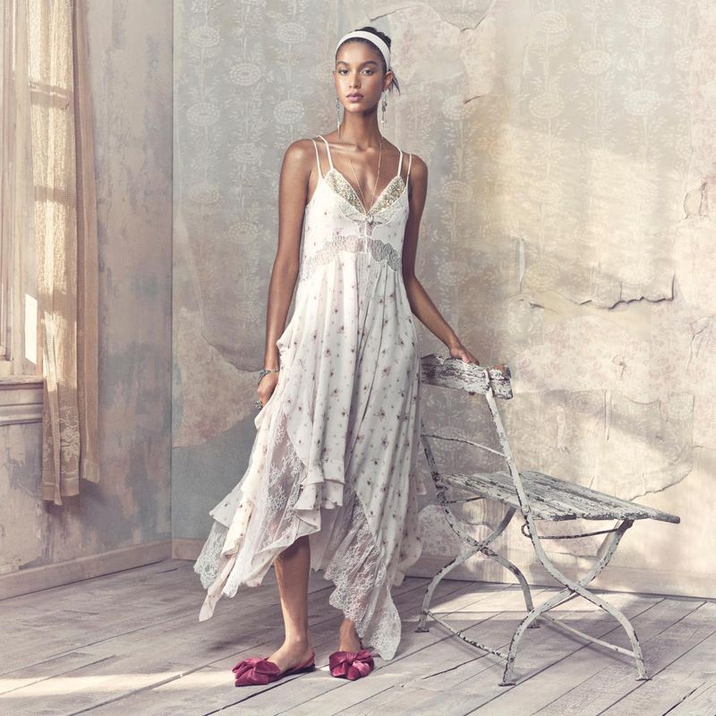 A woman wearing a patterned white dress with lace details and slides, standing next to an antique chair in an indoor studio setting.