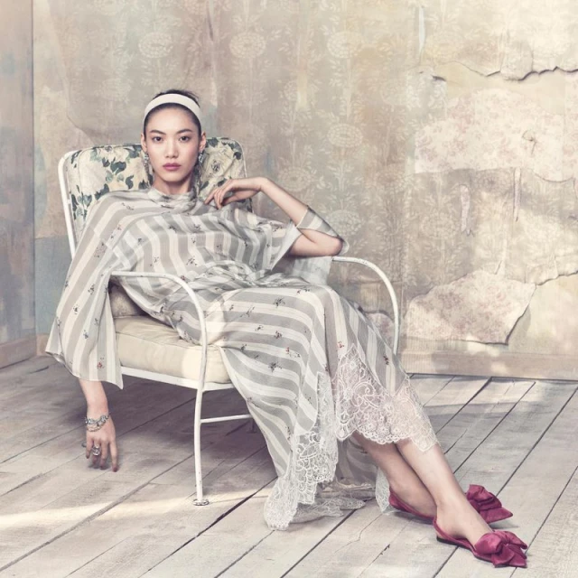 An adult woman sits in a chair wearing a striped dress and headband in an indoor setting with a weathered wall and wood floor.