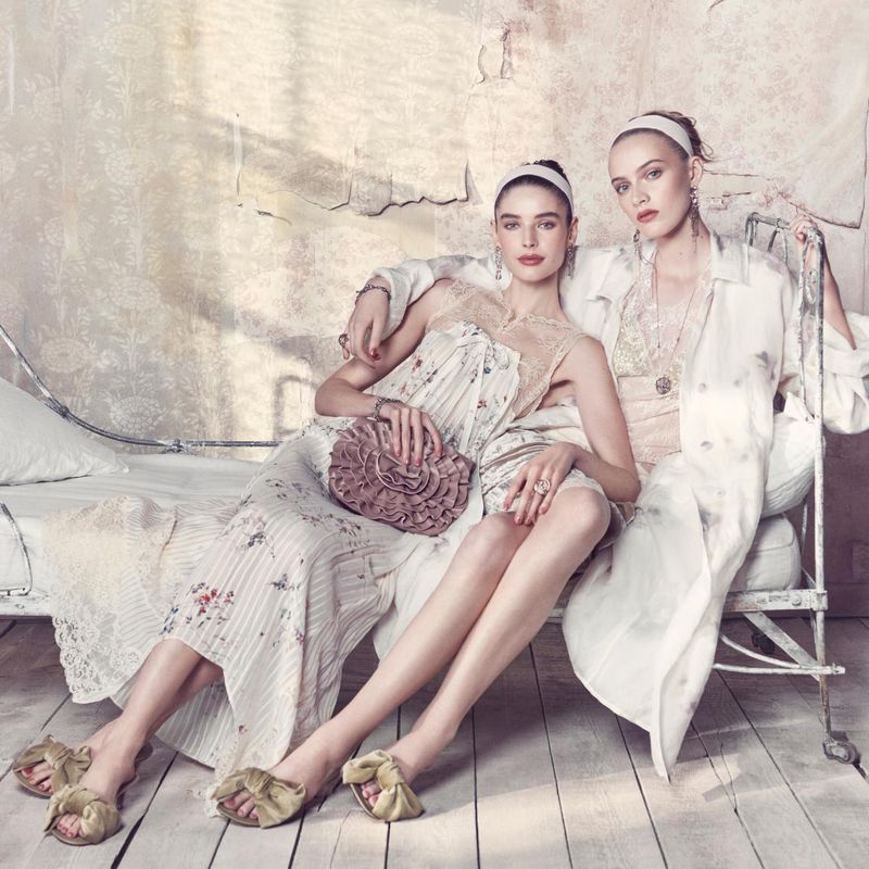 Two adult females in vintage-inspired floral dresses and headbands pose on a metal bed frame in a bright, indoor setting.
