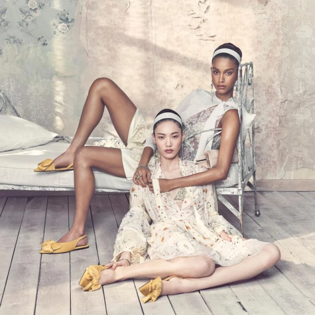 Two female models in an indoor setting. One reclines on a daybed wearing shorts and a headband, while the other sits on the floor.