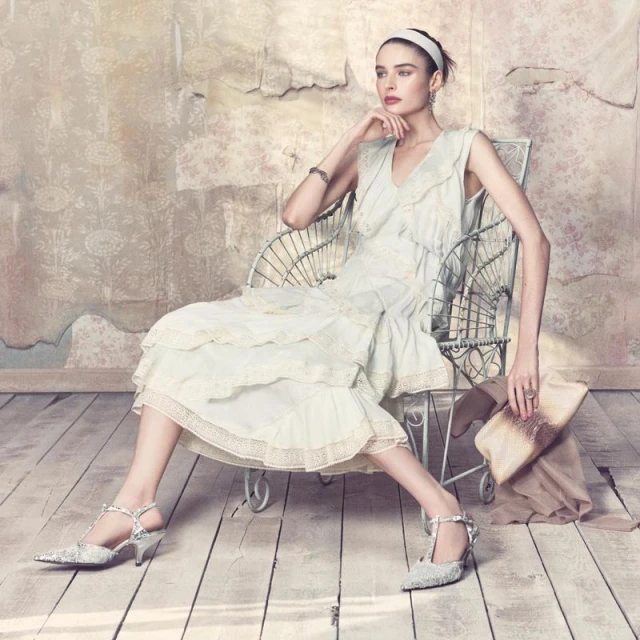 Elegant woman in vintage dress seated in wire chair with clutch and heels in a bright indoor studio setting with peeling wallpaper.