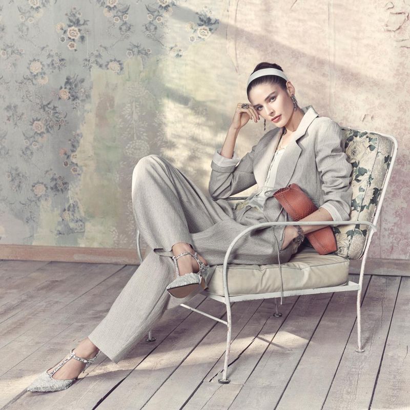 Elegant adult female posing in a light grey suit and embellished T-strap heels, seated in a floral-patterned chair against a floral wall.