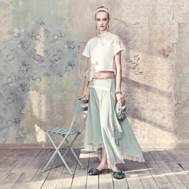 Fashion editorial shot: Woman in white top and mint skirt posing near a chair against floral wallpaper backdrop in soft, natural light.