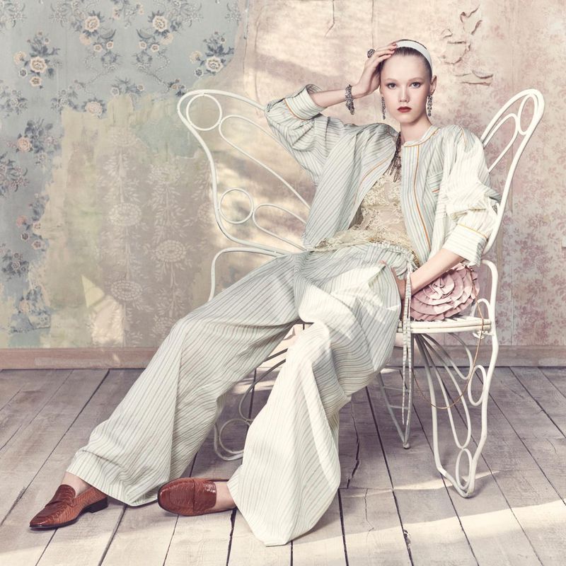 Fashionable woman in a striped suit sitting on a white butterfly chair in an indoor studio with a floral wallpaper backdrop.