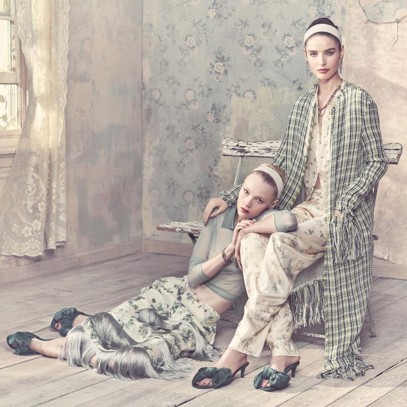 Two women in floral and plaid outfits pose indoors with vintage floral wallpaper, hardwood floors, and soft natural light from a window.