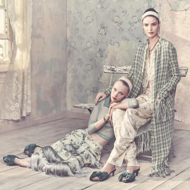 Two women in floral and plaid outfits pose indoors with vintage floral wallpaper, hardwood floors, and soft natural light from a window.