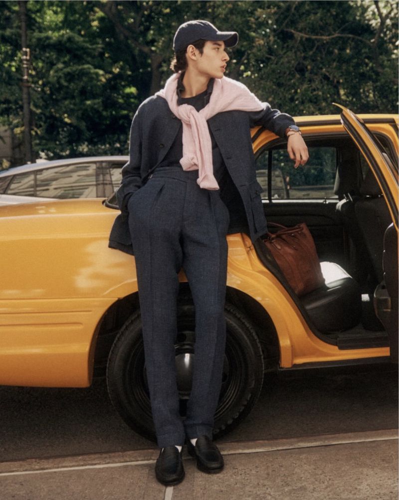 Young man in a dark blue jacket and pants stands beside a yellow taxi cab on a city street, wearing a baseball cap and loafers.