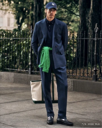 Man in suit and baseball cap standing on city sidewalk with tote bag, green sweater tied around shoulders. Outdoor fashion shot.