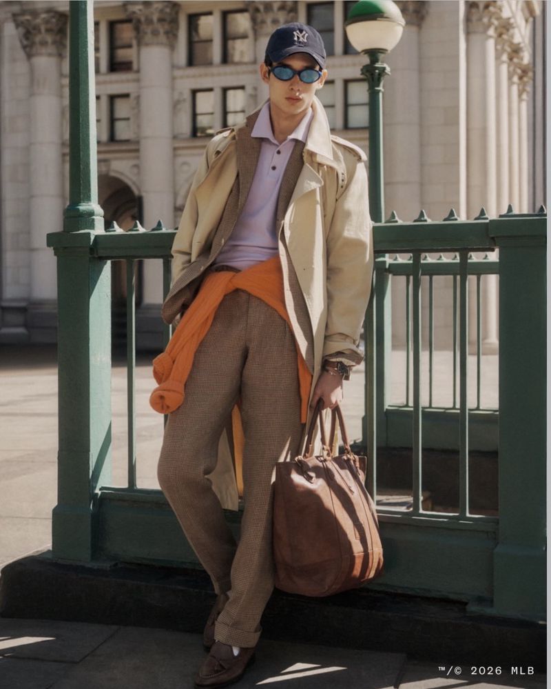 Stylish man in trench coat, baseball cap, and sunglasses poses in front of a building with an urban street lamp fixture.