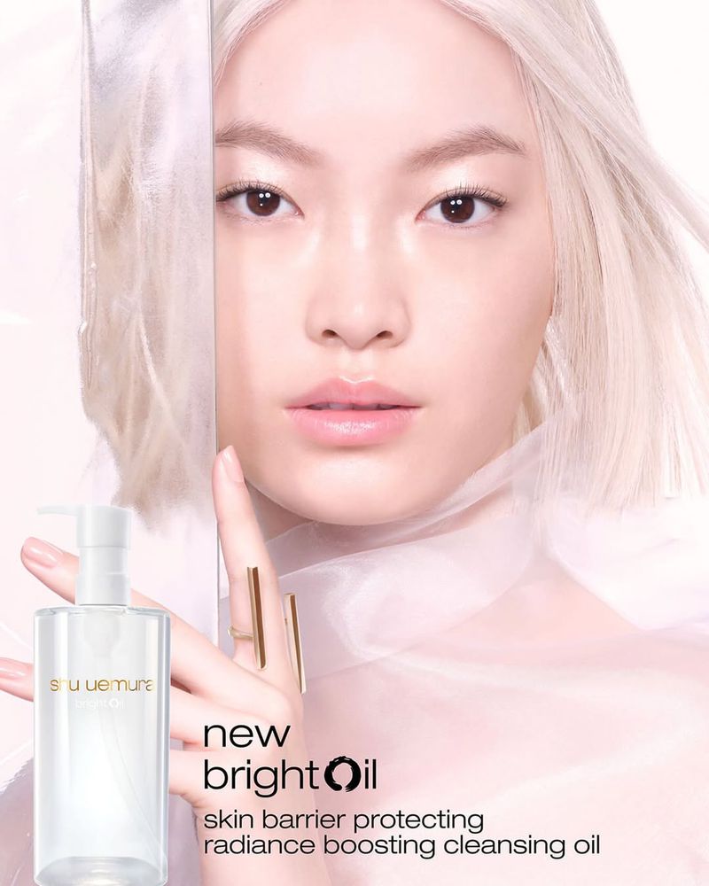 Shu Uemura skincare ad features an Asian woman with silver hair, clean skin, and light makeup, posing with a clear bottle of oil.
