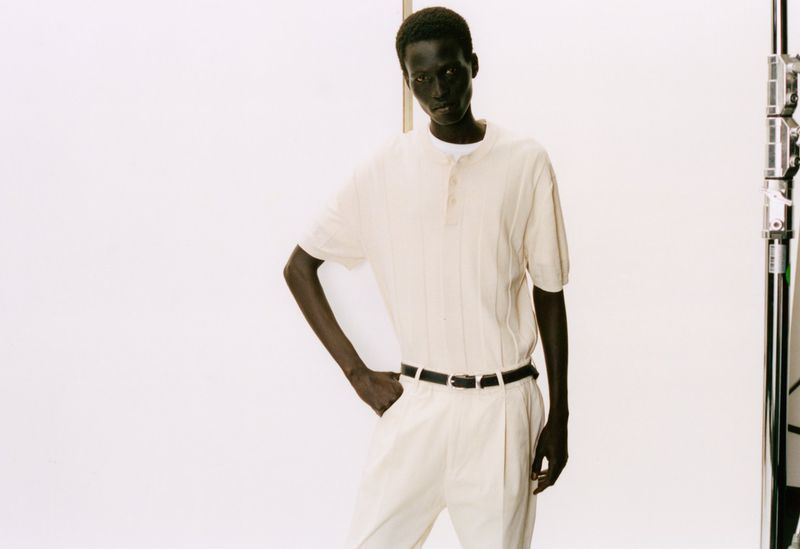 Studio fashion shot of a dark-skinned male model wearing a collared shirt and trousers, posing against a white backdrop with lighting equipment.
