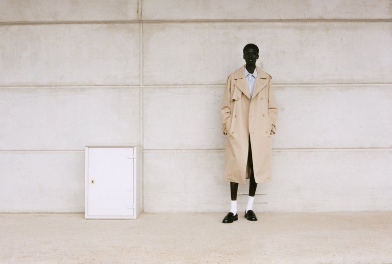 Fashionable adult man in trench coat posing near wall and white cabinet. The neutral palette creates a minimalist, modern aesthetic.