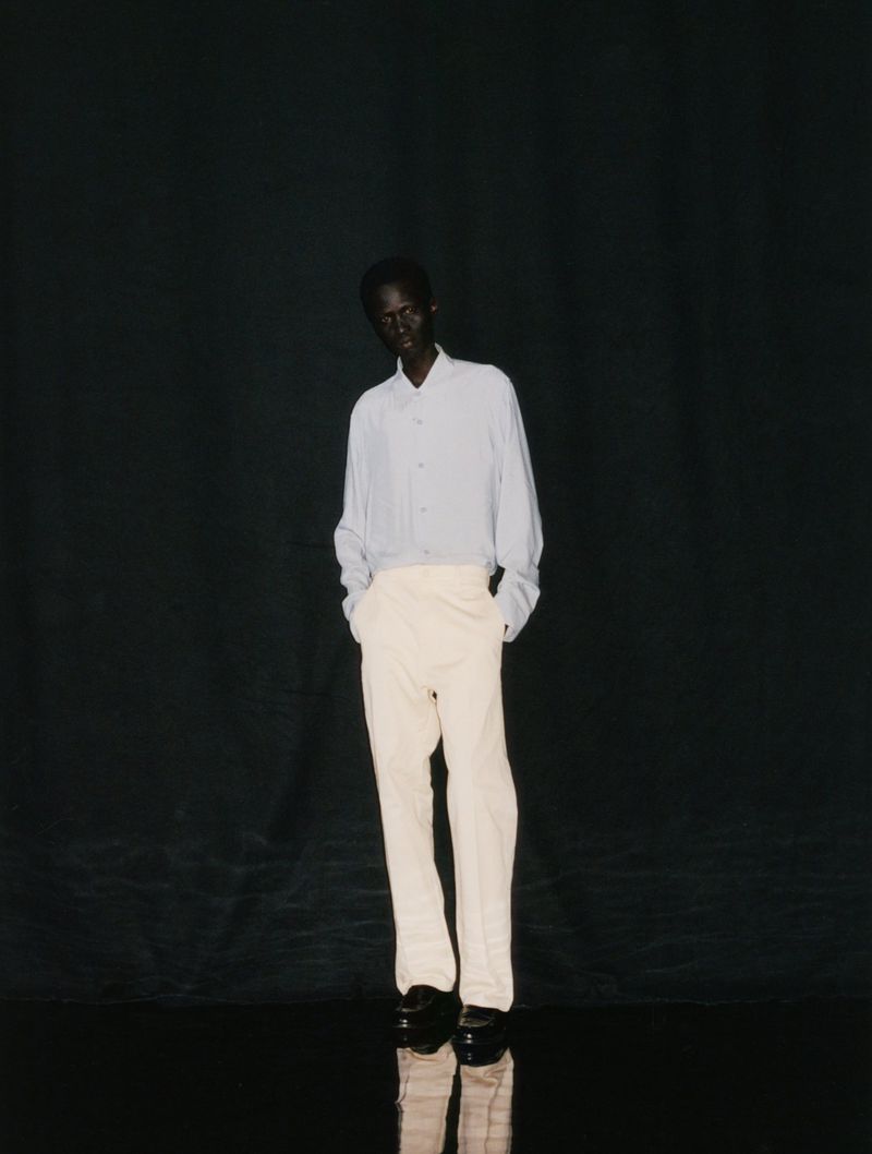 Dark-skinned male model stands in studio wearing light blue shirt and cream pants against a dark, reflective background.
