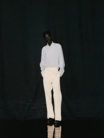 Dark-skinned male model stands in studio wearing light blue shirt and cream pants against a dark, reflective background.
