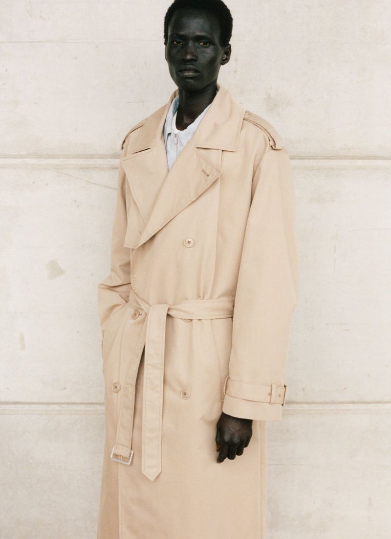 Adult male model with dark skin and a neutral expression poses in a tan trench coat in front of a textured off-white wall.