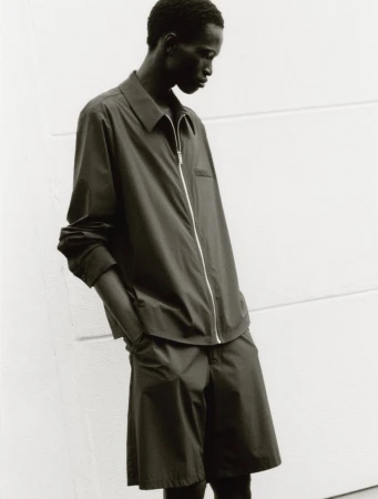 Monochromatic shot of an adult male model wearing a black button-down collared shirt and matching shorts in front of a plain white wall.