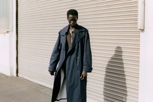 Model Mamuor Majeng in a dark navy trench coat posing outdoors against a beige ribbed roller shutter door.