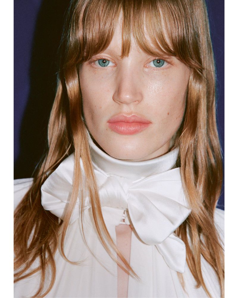 Close-up portrait of a fair-skinned woman with blonde hair, blue eyes, and a white top with a large bow, against a dark blue backdrop.