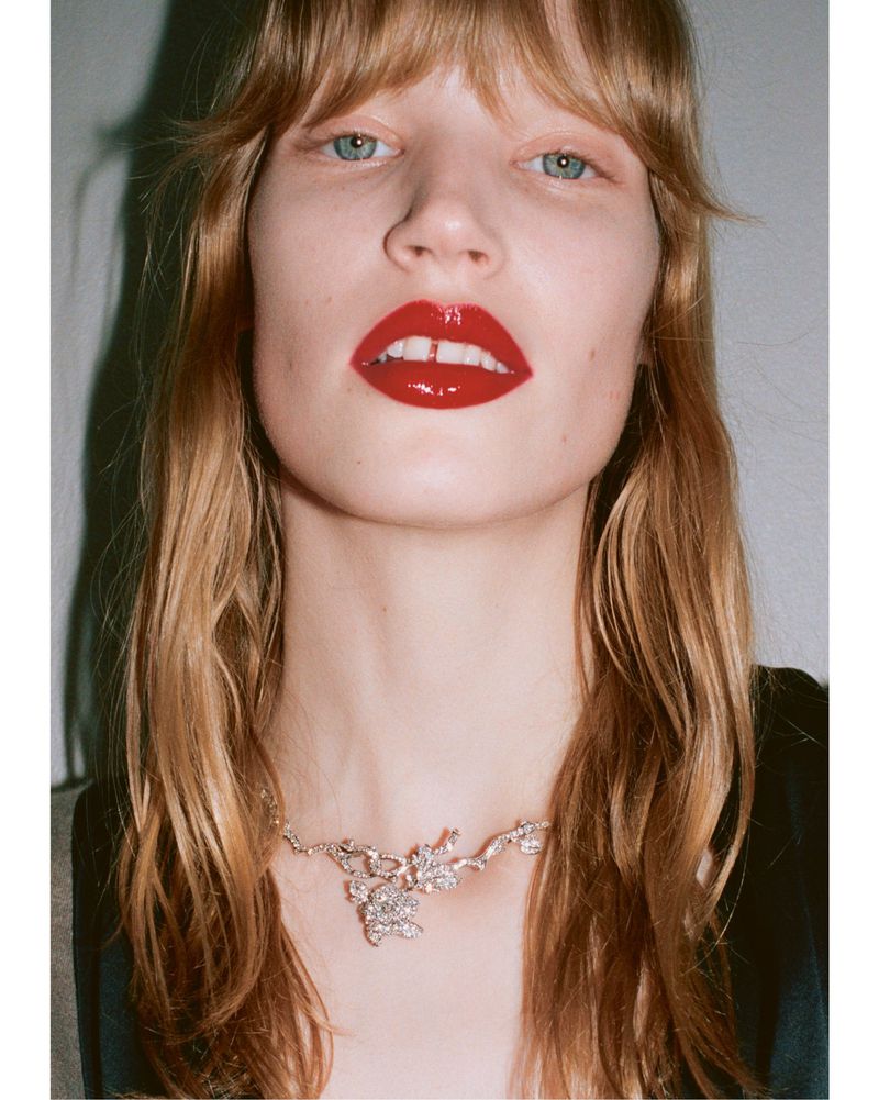 Close-up portrait of a woman with long red hair, glossy red lipstick, and a sparkling jeweled necklace.