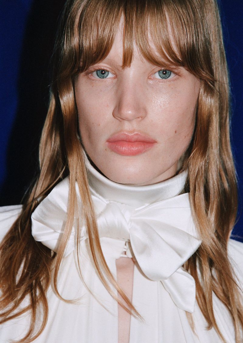 Close-up portrait of a fair-skinned adult woman with light brown hair and blue eyes wearing a white top and large bow against blue background.