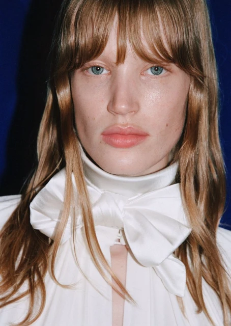 Close-up portrait of a fair-skinned adult woman with light brown hair and blue eyes wearing a white top and large bow against blue background.