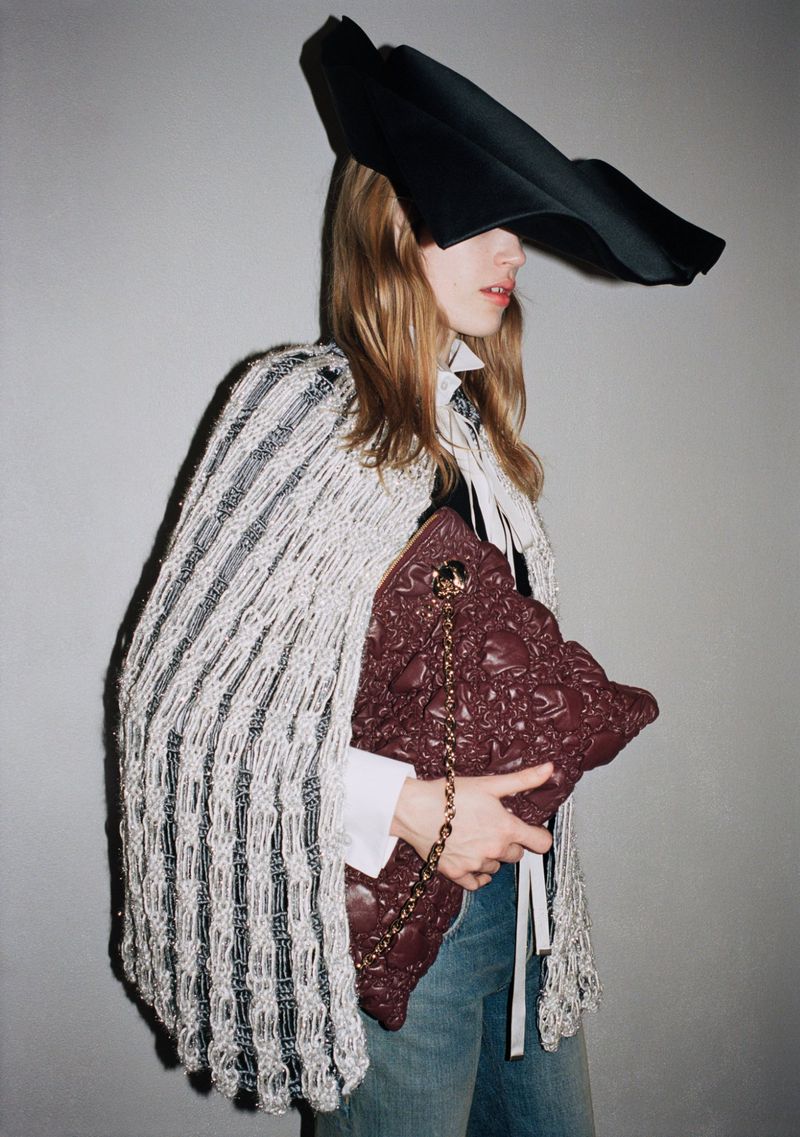 Model in wide black hat wearing textured white cape and holding maroon leather bag against grey backdrop