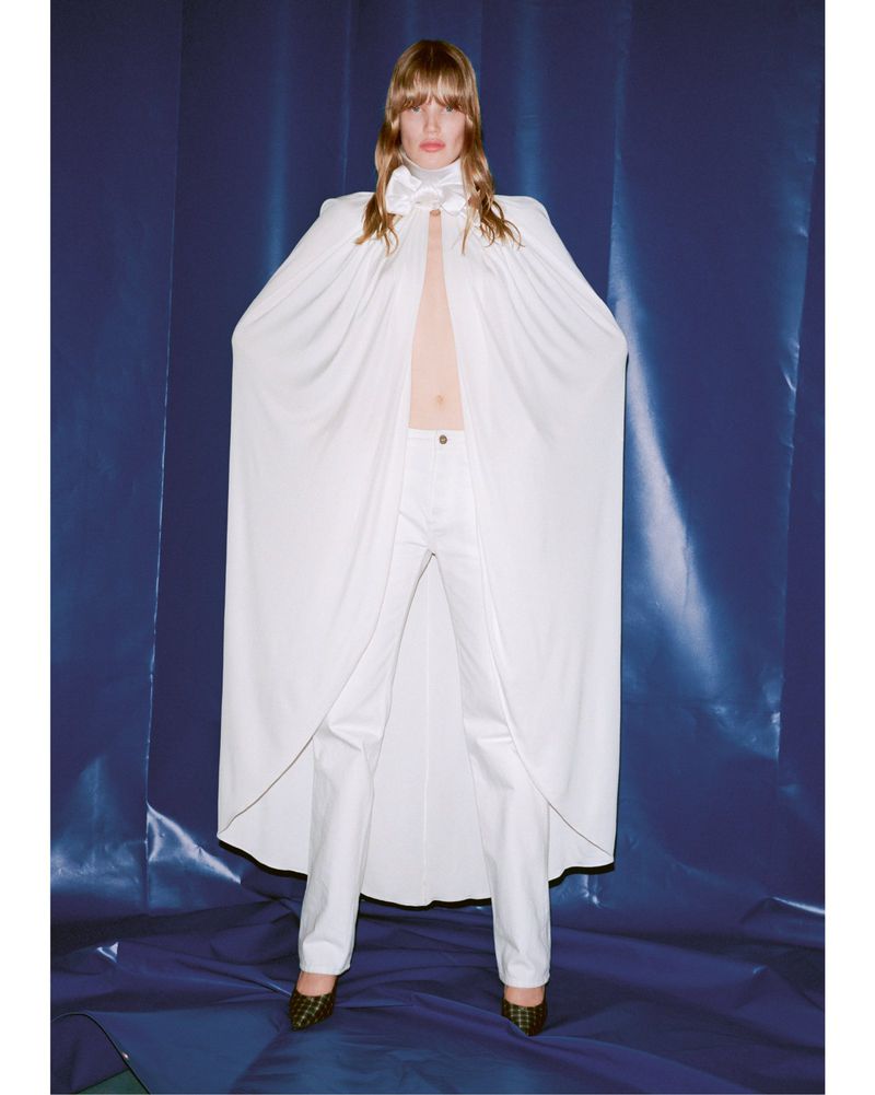 Full shot of a single fair-skinned fashion model in a white cape and white pants against a blue backdrop in a studio setting.