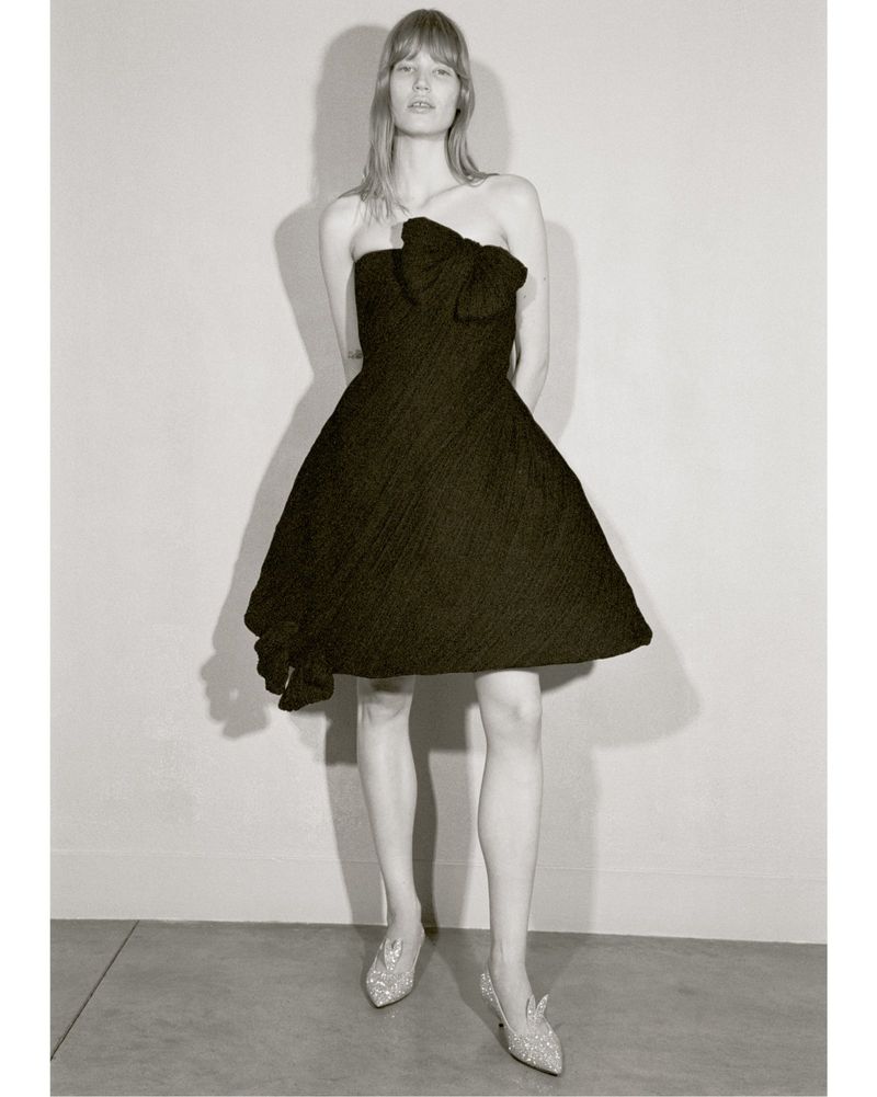 Elegant woman in a black strapless dress with a bow detail and silver heels, posing against a neutral background in a fashion editorial shot.