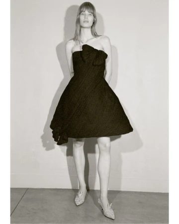 Elegant woman in a black strapless dress with a bow detail and silver heels, posing against a neutral background in a fashion editorial shot.