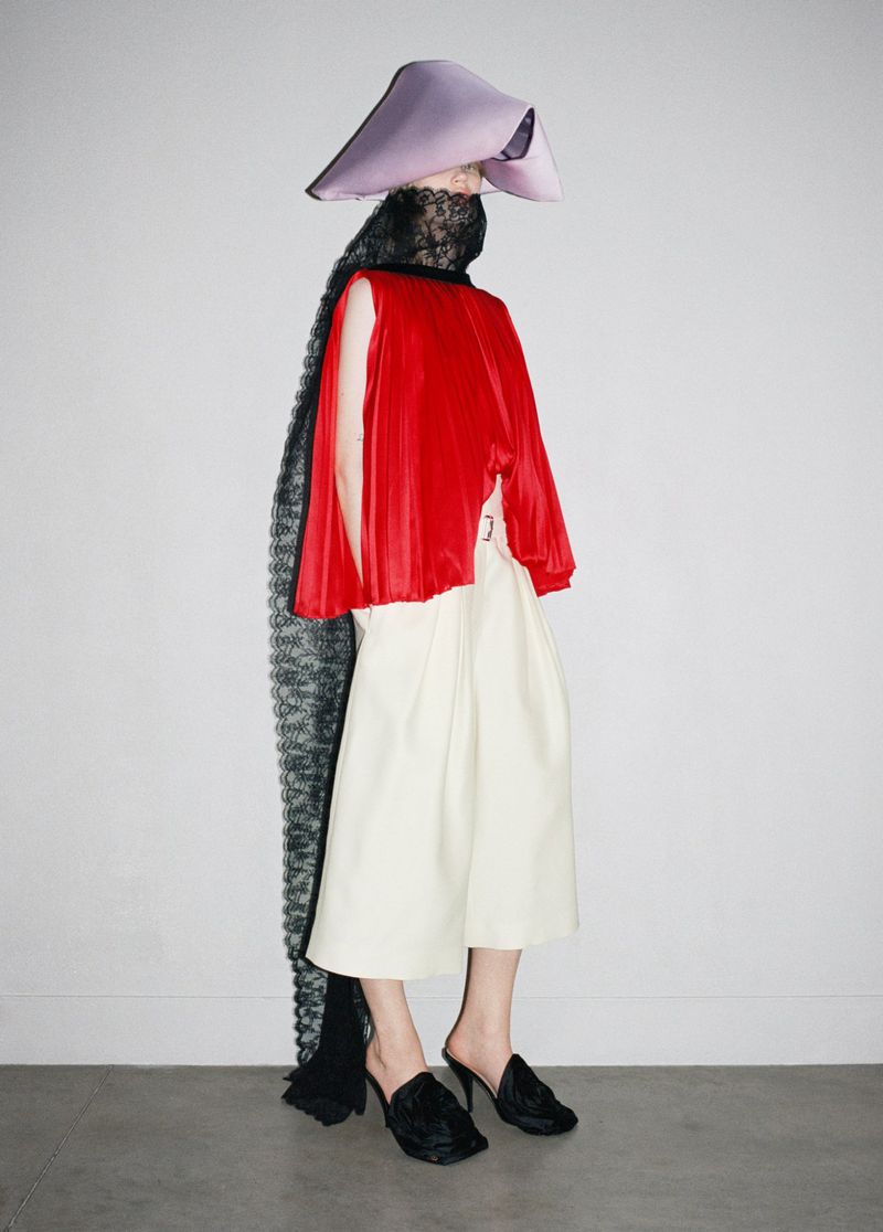 Fashion shot of a woman with lavender hat, red top, and black cape posing against white background in studio lighting, with black heels.