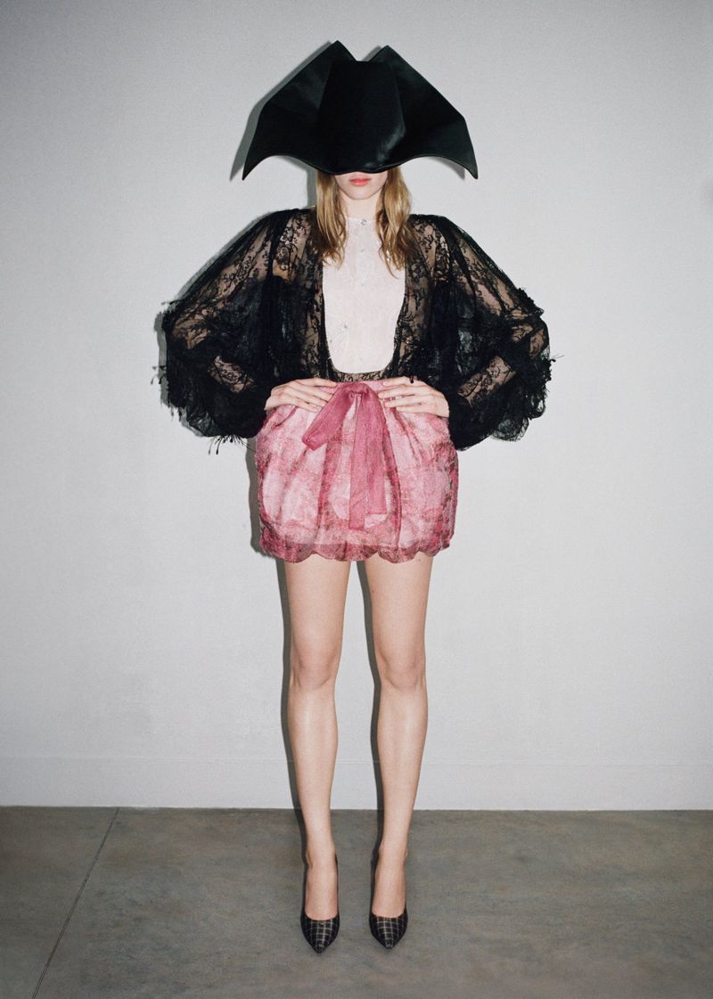 Model standing facing the camera wearing a sheer black lace top, pink bubble skirt, and an oversized black structural headpiece.