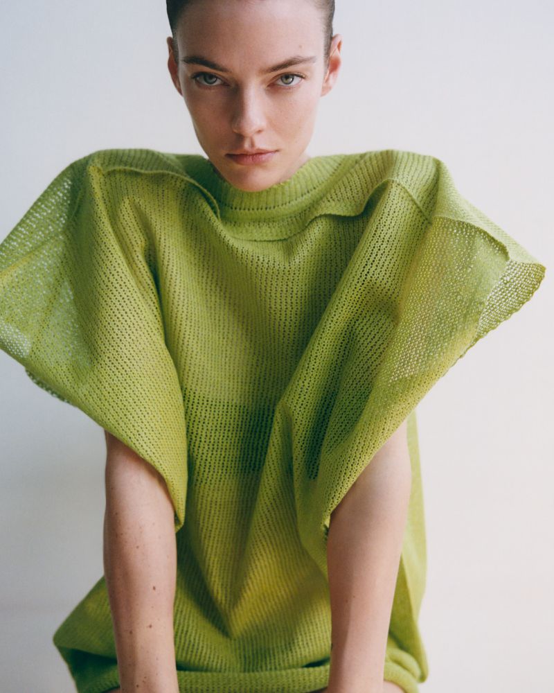 A woman with fair skin poses in a studio wearing a light green knit top; medium shot, neutral background, fashion advertisement.