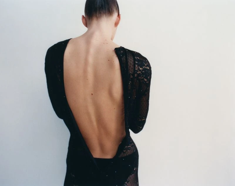 Back view of a woman with dark hair wearing a long-sleeved black lace dress with a plunging back, standing against a white wall.