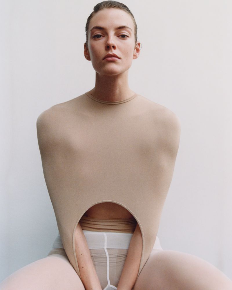 Woman in nude bodysuit with neutral makeup and sleek hairstyle, posing in a photo studio with a plain white background, looking at the camera.