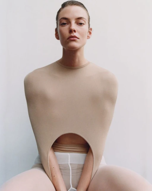 Woman in nude bodysuit with neutral makeup and sleek hairstyle, posing in a photo studio with a plain white background, looking at the camera.