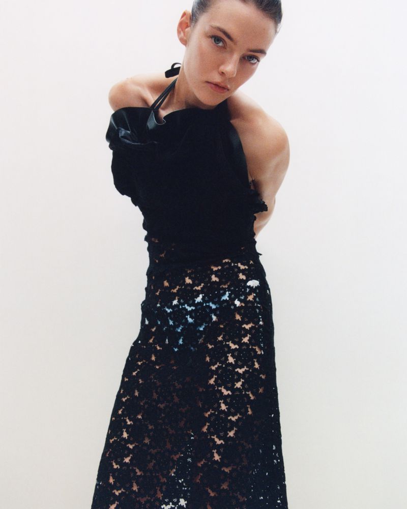 A fair-skinned woman poses in a black top and black lace skirt against a white background, in a fashion or editorial shot.