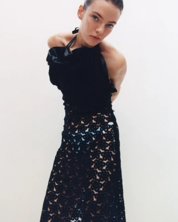 A fair-skinned woman poses in a black top and black lace skirt against a white background, in a fashion or editorial shot.