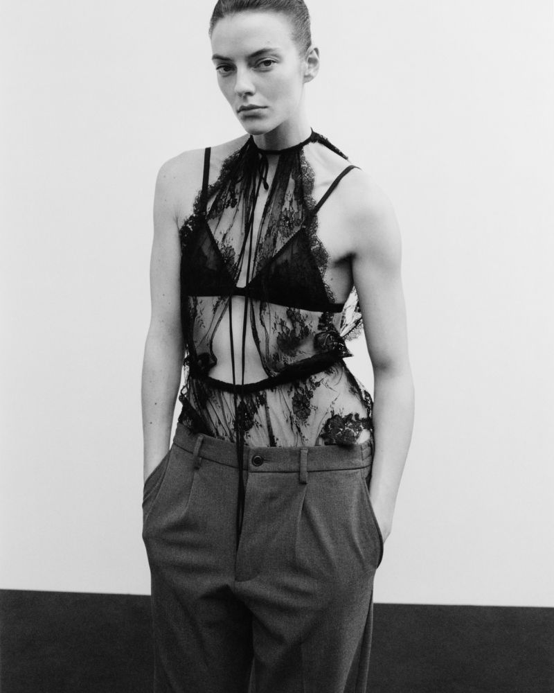 Black and white studio shot of a woman in a lacy black top and grey trousers with her hands in her pockets, looking at the camera.