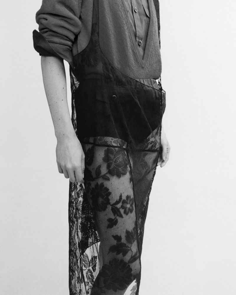 Black-and-white studio portrait of a person wearing a sheer, floral lace dress over a shirt and shorts against a plain backdrop.
