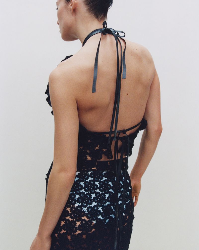 Adult woman poses in a black floral lace halter dress against a white background, photographed from the rear in a portrait orientation.