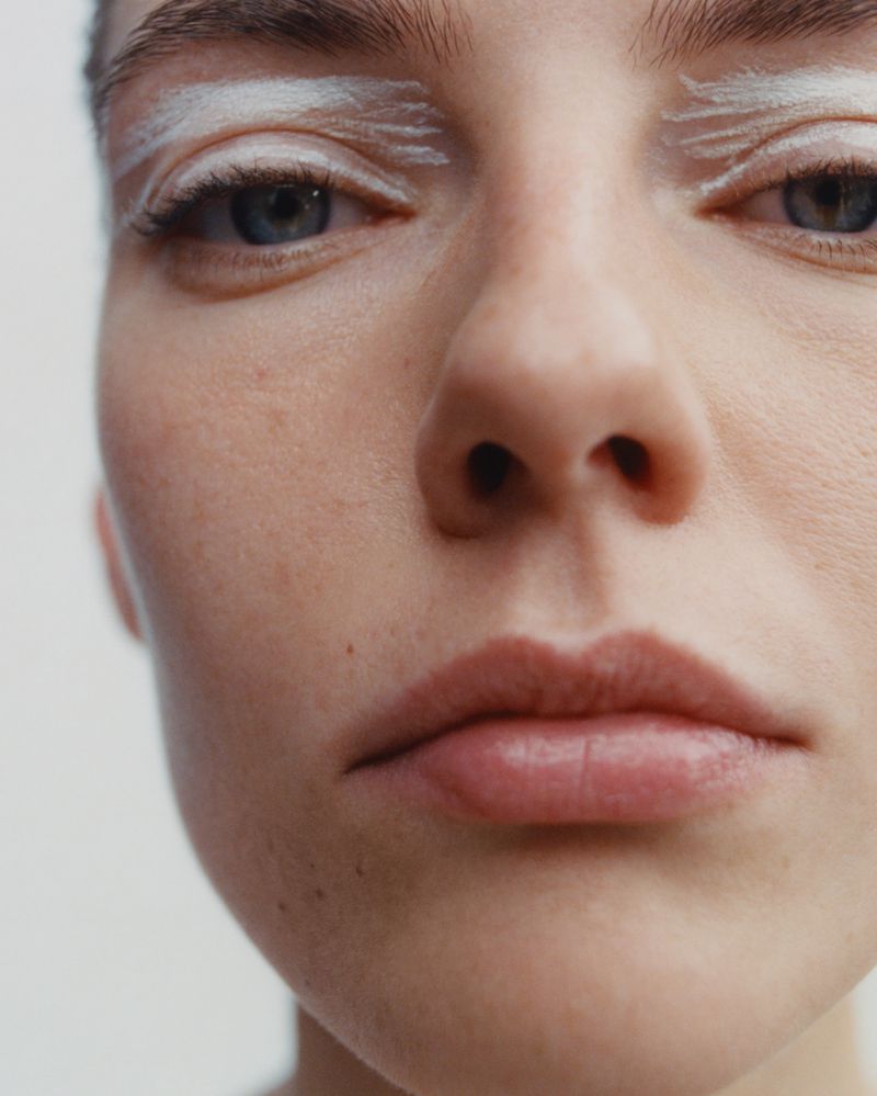 Close-up portrait of a young fair-skinned woman with white painted eyebrows, light blue eyes, and neutral expression against a white background.