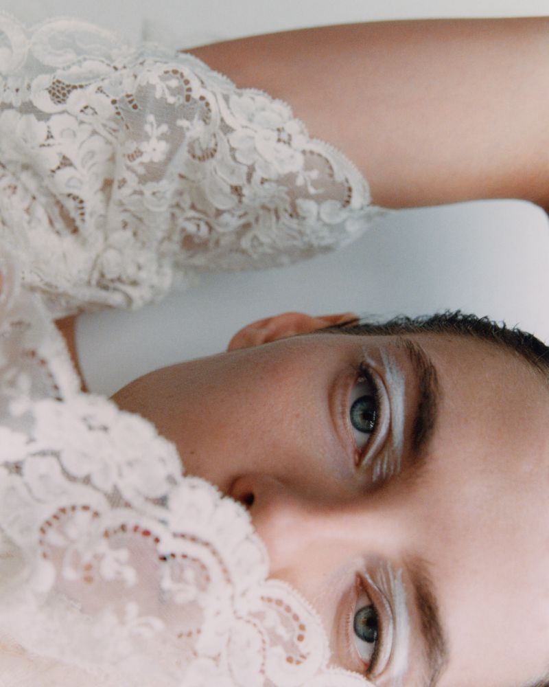 Close-up fashion portrait of a fair-skinned adult female with white eye makeup and blue eyes, wearing white lace against a white background.