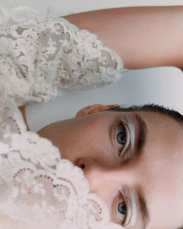 Close-up fashion portrait of a fair-skinned adult female with white eye makeup and blue eyes, wearing white lace against a white background.