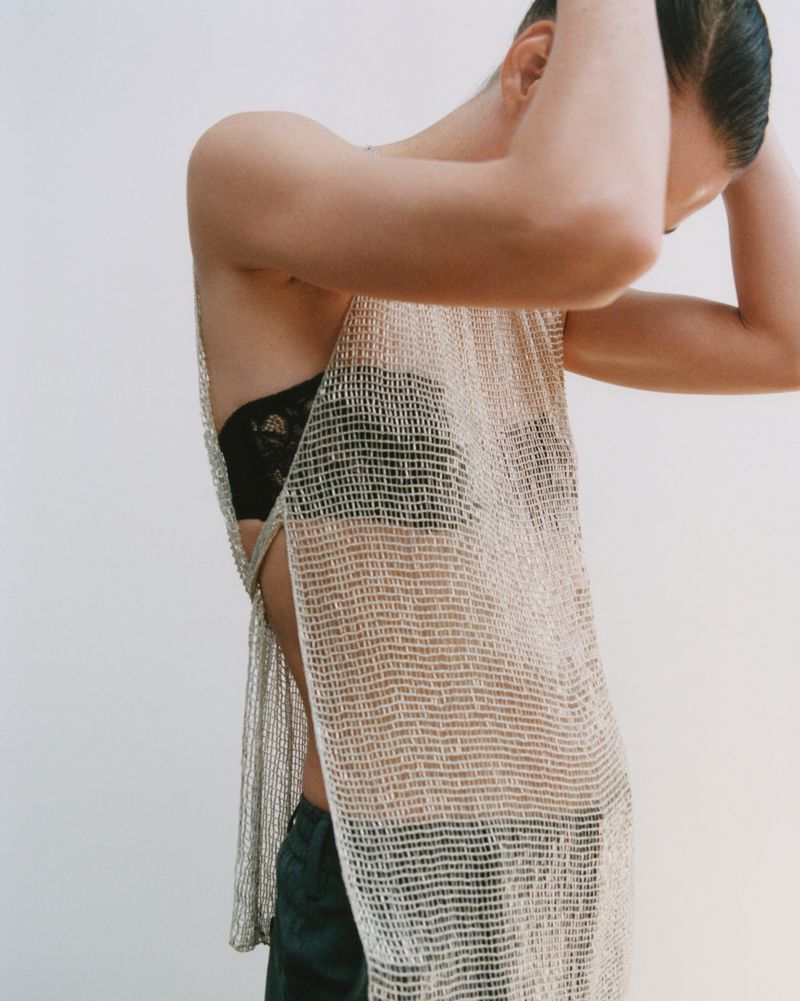 Woman in a shimmering, sheer silver dress and black bra poses in a studio with her hands behind her head. Minimalist beauty shot.