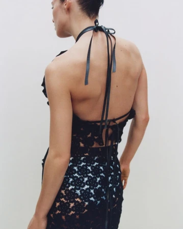 Model seen from behind wearing a black lace halterneck dress with thin leather straps against a neutral wall.