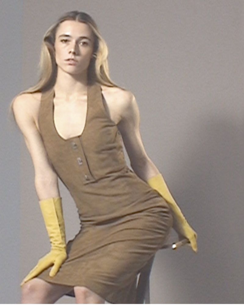 A person with long blonde hair, wearing a brown halter dress and yellow gloves, poses in a studio setting with a grey backdrop.